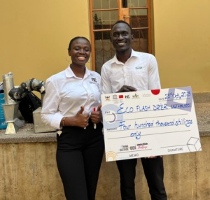 A photo of two people standing outdoors in front of a beige building with a tall wooden door behind them. On the right is Oscar. Both individuals are wearing light‑colored shirts with visible logos. One person is giving a thumbs‑up gesture. They are holding a large ceremonial cheque made out to “Eco Flash Dryer,” dated 23rd October 2025, for the amount of four hundred thousand shillings. Various equipment, including metallic cylindrical items, is visible on a ledge beside them.