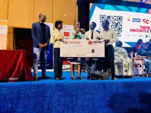 A photo taken on an indoor stage during an event, where five people stand together holding a large ceremonial cheque made out to “Eco Flash Dryer” for 1,500 units of currency. The stage has blue carpeting, and event banners and a large screen displaying a QR code and the text “Youth Skills Fest” are visible in the background.